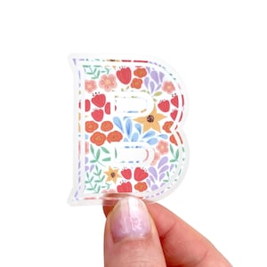 May include: A clear sticker in the shape of the letter "B", decorated with a floral pattern. The design includes red, orange, yellow, and blue flowers on a white background. The sticker is held by a hand.
