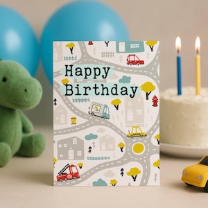 May include: A birthday card with a road map design featuring cars, trees, and houses. The card reads "Happy Birthday" in large, bold letters. A toy car and a cake with lit candles are also in the image.