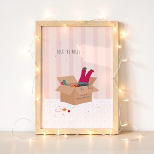 May include: A framed print with the text "Deck the Halls." The artwork depicts a person's legs sticking out of a cardboard box filled with colorful decorations. The box is labeled "xmas decorations" and sits on a pink and white striped background.