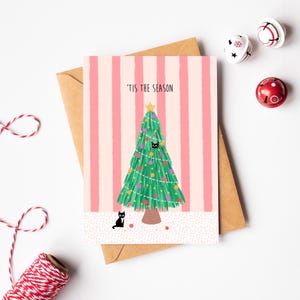 Könnte beinhalten: Weihnachtskarte mit einem grünen Weihnachtsbaum, verziert mit bunten Kugeln und einem goldenen Stern. Die Karte trägt den Text "'TIS THE SEASON" und zeigt eine schwarze Katze. Die Karte liegt auf einem braunen Umschlag.