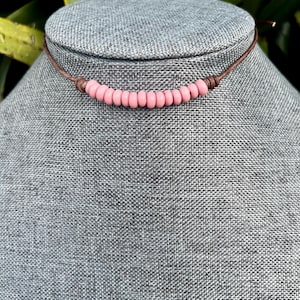 May include: A pink beaded choker necklace. The necklace features small, round, light pink beads strung together on a brown leather cord. The cord is knotted at each end. The necklace is displayed on a gray mannequin.