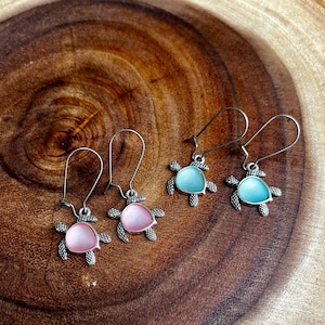 May include: Three pairs of silver turtle earrings with iridescent blue and pink accents. The earrings are on a wooden surface.