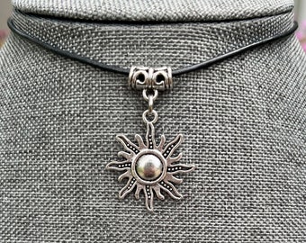 May include: A silver sun pendant necklace with a black leather cord. The sun pendant has a detailed design with rays emanating from the center.