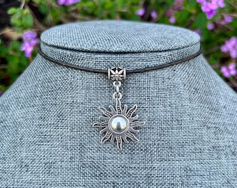 May include: A silver sun pendant necklace on a black leather cord. The pendant features a detailed sun design with a round center.