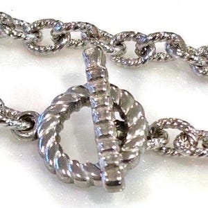 May include: Close-up of a silver-colored necklace with a textured chain and a toggle clasp. The chain links are oval-shaped and twisted. The clasp is a circular design with a bar.