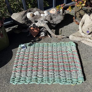 Puede incluir: Una alfombra rectangular hecha de cuerda tejida en tonos de verde, rosa y blanco. La alfombra tiene una superficie texturizada y un borde festoneado. La alfombra está colocada sobre una superficie de hormigón gris.