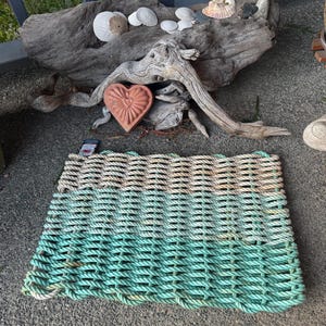 Puede incluir: Una alfombra rectangular hecha de cuerda tejida en tres secciones horizontales: verde, beige y blanquecino. Un tronco de deriva con conchas marinas y un adorno en forma de corazón están en el fondo. La alfombra está sobre una superficie de hormigón.