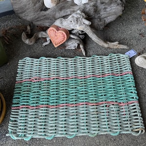 Puede incluir: Una alfombra rectangular hecha de cuerda tejida en tonos verde mar y gris, con una franja horizontal de cuerda rosa. La alfombra está colocada sobre una superficie de hormigón gris, con madera flotante y conchas marinas en el fondo.