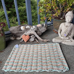 Puede incluir: Una alfombra rectangular hecha de cuerda tejida en filas alternas de colores azul claro y melocotón. La alfombra está colocada sobre una superficie de hormigón, con una estatua de Buda y una planta en maceta en el fondo.