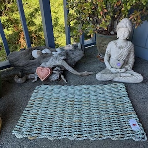 Puede incluir: Una alfombra rectangular de cuerda trenzada azul claro con un borde festoneado. La alfombra se exhibe al aire libre con una estatua de Buda, un objeto en forma de corazón y plantas en macetas en el fondo. La alfombra mide aproximadamente 61 cm de largo y 41 cm de ancho.