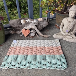 Puede incluir: Una alfombra rectangular hecha de cuerda tejida. La alfombra está dividida en tres secciones horizontales: blanco, coral y verde agua. La alfombra está colocada sobre una superficie de hormigón, con un fondo borroso de vegetación y una estatua de Buda.