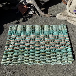 Puede incluir: Una alfombra rectangular tejida con cuerda gruesa y duradera. La alfombra presenta rayas horizontales en verde mar, rosa claro y verde más oscuro. Los bordes tienen un diseño festoneado, agregando un toque decorativo.