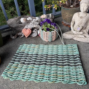 Puede incluir: Una alfombra rectangular hecha de cuerda tejida en tonos verde mar, negro y gris. La alfombra tiene un borde festoneado y está colocada sobre una superficie de hormigón. La alfombra es un elemento decorativo para una entrada.