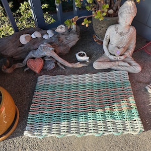 May include: A rectangular woven rope mat featuring horizontal stripes in alternating shades of light green, soft pink, and off-white. The mat is constructed from thick, textured cordage arranged in a tight, repeating grid pattern with finished edges.
