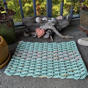 Puede incluir: Una alfombra rectangular tejida con cuerda verde agua con un borde festoneado. La alfombra presenta dos rayas horizontales de cuerda rosa y blanca. La alfombra está colocada sobre una superficie gris.