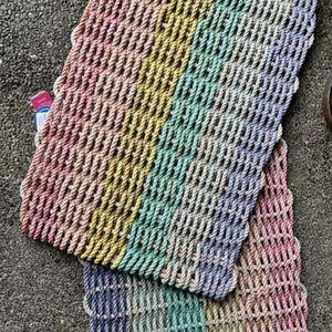 Puede incluir: Dos felpudos rectangulares hechos de cuerda trenzada. Las alfombras están divididas en franjas verticales de color, incluyendo rosa, amarillo, verde y azul. Las alfombras están colocadas sobre una superficie gris.