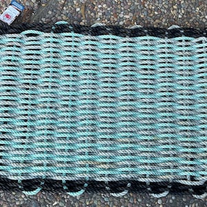 Puede incluir: Felpudo rectangular tejido con cuerda gruesa en tonos azules, grises y negros. La alfombra tiene una superficie texturizada y un borde de cuerda negra. Una pequeña etiqueta es visible en la esquina superior izquierda.
