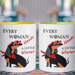 May include: Two white ceramic mugs with yellow and green rims. Both mugs feature a black and brown dachshund wearing heart-shaped sunglasses and a red bandana. The text on the mugs reads "Every Woman Needs A Little Wiener".