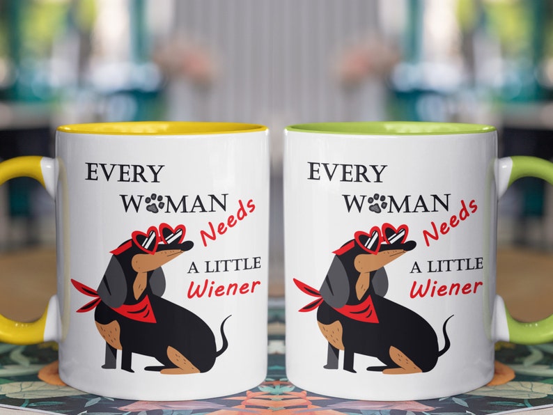 May include: Two white ceramic mugs with yellow and green rims. Both mugs feature a black and brown dachshund wearing heart-shaped sunglasses and a red bandana. The text on the mugs reads "Every Woman Needs A Little Wiener".