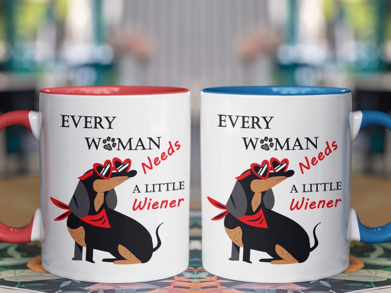 May include: Two white ceramic mugs with red and blue rims. Both mugs feature a black dachshund wearing red heart-shaped sunglasses and a red bandana. The text on the mugs reads "Every Woman Needs A Little Wiener".