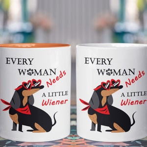May include: Two white ceramic mugs with orange handles. The mugs feature a black and brown dachshund wearing red heart-shaped sunglasses and a red bandana. The text on the mugs reads "Every Woman Needs A Little Wiener".