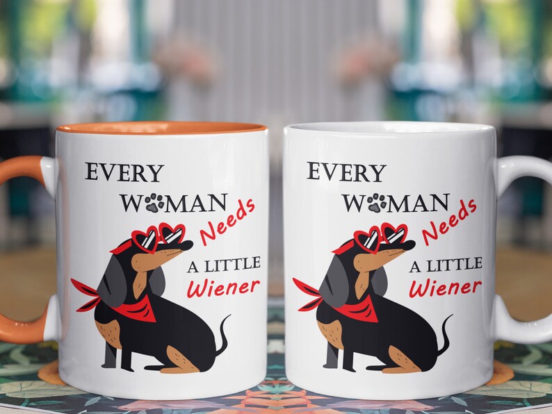 May include: Two white ceramic mugs with orange handles. The mugs feature a black and brown dachshund wearing red heart-shaped sunglasses and a red bandana. The text on the mugs reads "Every Woman Needs A Little Wiener".