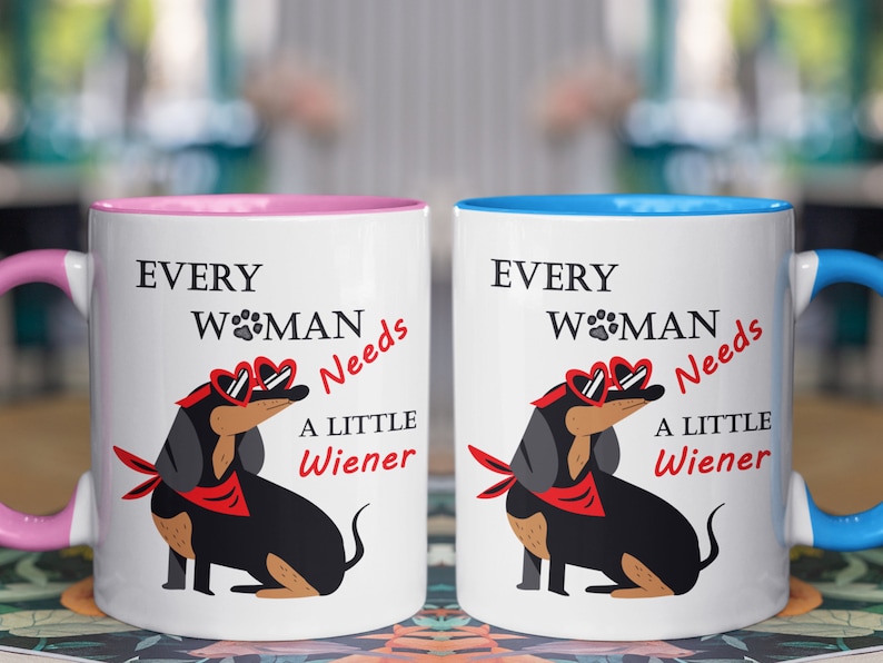 May include: Two white ceramic mugs with pink and blue rims. Each mug features a black dachshund wearing red heart-shaped sunglasses and a red bandana. The text on the mugs reads "Every Woman Needs A Little Wiener".