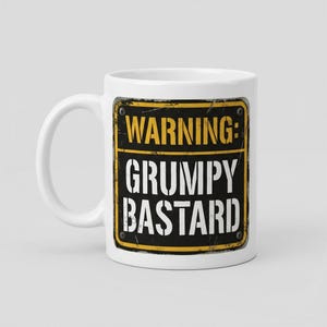 Op de afbeelding: Een witte keramische mok met een rechthoekige zwart-gele waarschuwingsbordafbeelding. De tekst luidt WARNING: GRUMPY BASTARD in vetgedrukte, verweerde sjabloonletters. Het ontwerp bevat vier kleine schroefkoppen op de hoeken van het bord.