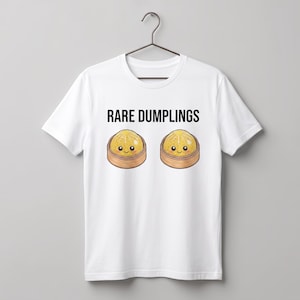 May include: A white short-sleeved t-shirt featuring the black text RARE DUMPLINGS above two illustrations of yellow dumplings with smiling faces, each sitting inside a small brown bamboo steamer basket, displayed on a dark metal hanger.