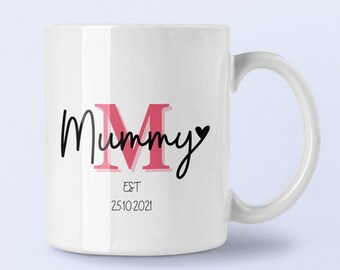 Puede incluir: Una taza de cerámica blanca con el texto Mummy en negro, una letra M grande de color rosa en el centro y un pequeño corazón negro al final. Debajo del texto, las palabras EST y la fecha 25.10.2021 están impresas en una fuente negra sencilla.