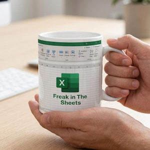 May include: A white ceramic mug featuring a printed graphic of a spreadsheet interface with the Microsoft Excel logo and the text "Freak in The Sheets" in dark green lettering. The design includes a ribbon menu at the top and a grid pattern.