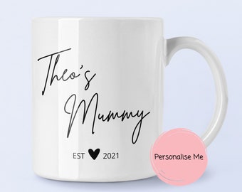Puede incluir: Una taza de cerámica blanca con texto en cursiva negra que dice Theo's Mummy, junto con el texto EST 2021 separado por un pequeño icono de corazón negro. La taza tiene un acabado liso y brillante con un asa lateral estándar.