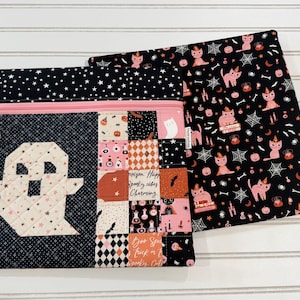 May include: Two quilted fabric pouches with zipper closures. One features a ghost appliqué on a black star-patterned background next to a patchwork section with text including 'Poison', 'Happy', 'Spooky vibes', and 'Charming'. The other has a black cat print.