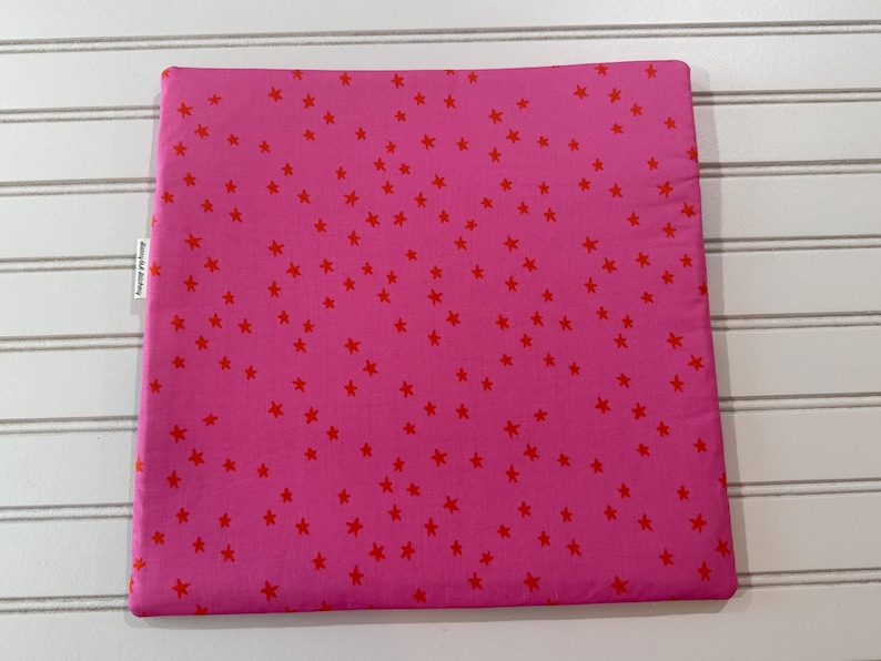 Peut inclure: Une housse de coussin carr&eacute;e en tissu rose vif, orn&eacute;e d'un motif dispers&eacute; de petites &eacute;toiles rouges pleines sur toute la surface. Une petite &eacute;tiquette en tissu blanc avec du texte noir est fix&eacute;e sur le bord lat&eacute;ral gauche de l'article textile.