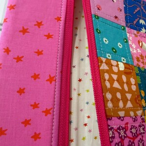 Peut inclure: Une pochette zipp&eacute;e avec un panneau en tissu rose &agrave; &eacute;toiles orange sur la gauche et un panneau en patchwork multicolore sur la droite. Le patchwork comprend des carr&eacute;s &agrave; motifs floraux, g&eacute;om&eacute;triques et d'insectes. Un tissu cr&egrave;me &agrave; petites &eacute;toiles est visible &agrave; l'int&eacute;rieur.
