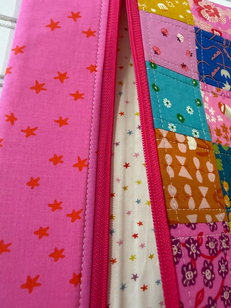 Peut inclure: Une pochette zipp&eacute;e avec un panneau en tissu rose &agrave; &eacute;toiles orange sur la gauche et un panneau en patchwork multicolore sur la droite. Le patchwork comprend des carr&eacute;s &agrave; motifs floraux, g&eacute;om&eacute;triques et d'insectes. Un tissu cr&egrave;me &agrave; petites &eacute;toiles est visible &agrave; l'int&eacute;rieur.
