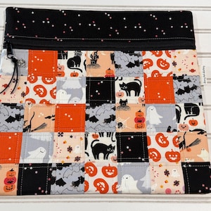 May include: A square zippered pouch featuring a patchwork design of Halloween-themed fabric squares. The pattern includes black cats, ghosts, pumpkins, bats, and spiders in orange, black, white, and gray. A black zipper with a small charm is at the top.