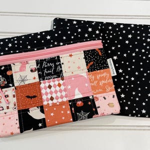 May include: A rectangular zippered pouch featuring a patchwork design of Halloween-themed fabric squares in black, orange, and pink, with a black fabric top and back printed with white stars. The pouch has a light pink zipper and a small charm attached.