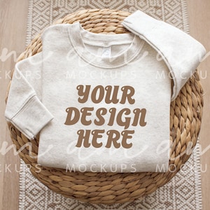 May include: A light gray sweatshirt with the text "YOUR DESIGN HERE" in brown, displayed on a woven basket. The sweatshirt is folded neatly, showcasing its design. The background includes a patterned rug.