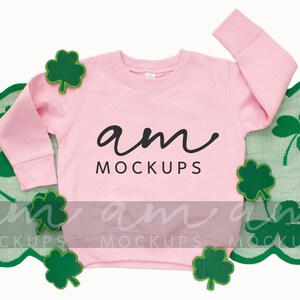 Puede incluir: Sudadera rosa de manga larga con el texto "am mockups" en la parte delantera. La sudadera está rodeada de decoraciones verdes en forma de trébol.