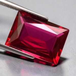 May include: A rectangular faceted gemstone with a vibrant, deep red color. The stone is held by a pair of silver-toned metal tweezers, showcasing its transparent clarity and precise geometric cut with multiple angled facets reflecting light.