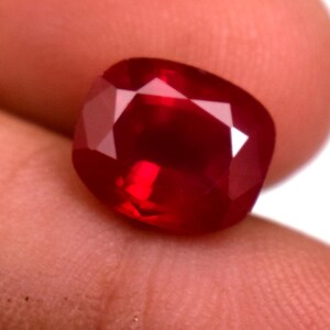 Puede incluir: Una gema de rubí tallada en cojín y facetada. La gema es de un color rojo intenso y saturado con múltiples facetas que reflejan la luz. La forma es un cuadrado redondeado, mostrando el brillo de la piedra.