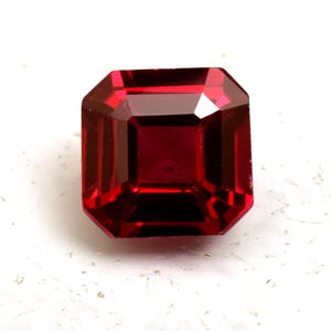 Puede incluir: Una gema roja oscura tallada en forma rectangular. La gema tiene forma cuadrada con bordes biselados, reflejando la luz y mostrando su rico color. La piedra está sobre un fondo blanco.