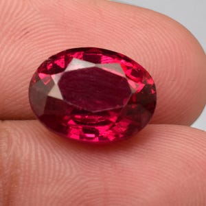 May include: A vibrant, oval-cut gemstone with a deep, saturated red color and multiple facets that reflect light. The stone features a polished surface with a clear, transparent quality, showcasing a brilliant crimson hue throughout its structure.