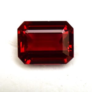 May include: A rectangular-cut, deep red gemstone with multiple facets. The gem has a brilliant, reflective surface with sharp, angled edges. The stone is set against a white background, highlighting its rich colour and clarity.