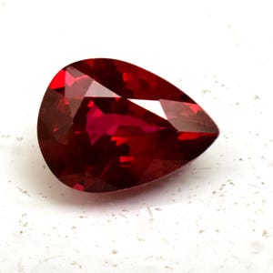 Op de afbeelding: Een gefacetteerde, peervormige edelsteen met een dieprode kleur. Het oppervlak van de edelsteen reflecteert licht en toont de vele facetten en de interne structuur. De steen is geplaatst tegen een witte achtergrond, wat de kleur en vorm benadrukt. Deze edelsteen is geschikt voor sieraden.