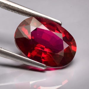 May include: An oval-cut, deep red gemstone with multiple facets that reflect light, held by a pair of silver-toned metal tweezers. The stone displays a vibrant, saturated crimson hue with internal clarity and sharp, geometric edges throughout.