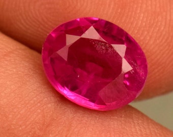 May include: A vibrant pink oval-shaped gemstone with a faceted cut. The gemstone is set against a neutral background.