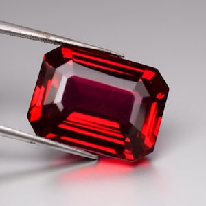 Puede incluir: Una gema facetada de corte rectangular sostenida por unas pinzas. La gema es de un color rojo intenso con un interior ligeramente más oscuro. Las facetas crean un juego de luces y sombras, realzando su brillo. Es probable que sea un rubí o un granate.