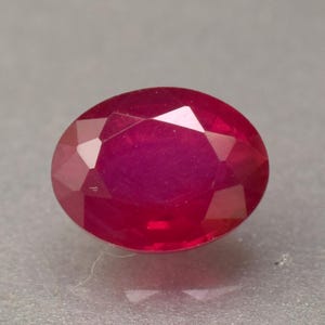 May include: A faceted, oval-cut ruby gemstone. The gem is a deep, saturated red colour with multiple reflective facets. The shape is elongated, with a polished surface that catches the light.